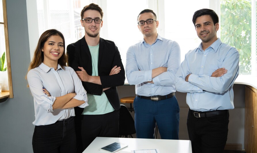Positive successful business team posing at workplace. Group of four managers standing at meeting table with arms crossed. Successful teamwork concept